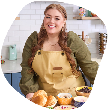 Smiling woman in an apron stands in a kitchen, leaning over a countertop with an assortment of ingredients. 